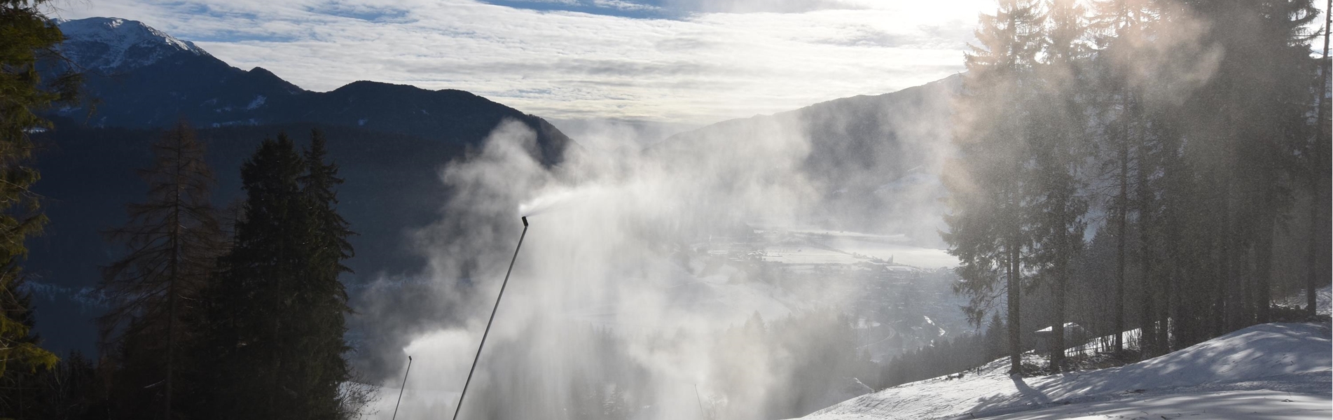 Schneebedeckte Skipiste mit Schneekanonen, Bäumen und Berglandschaft im Gegenlicht.