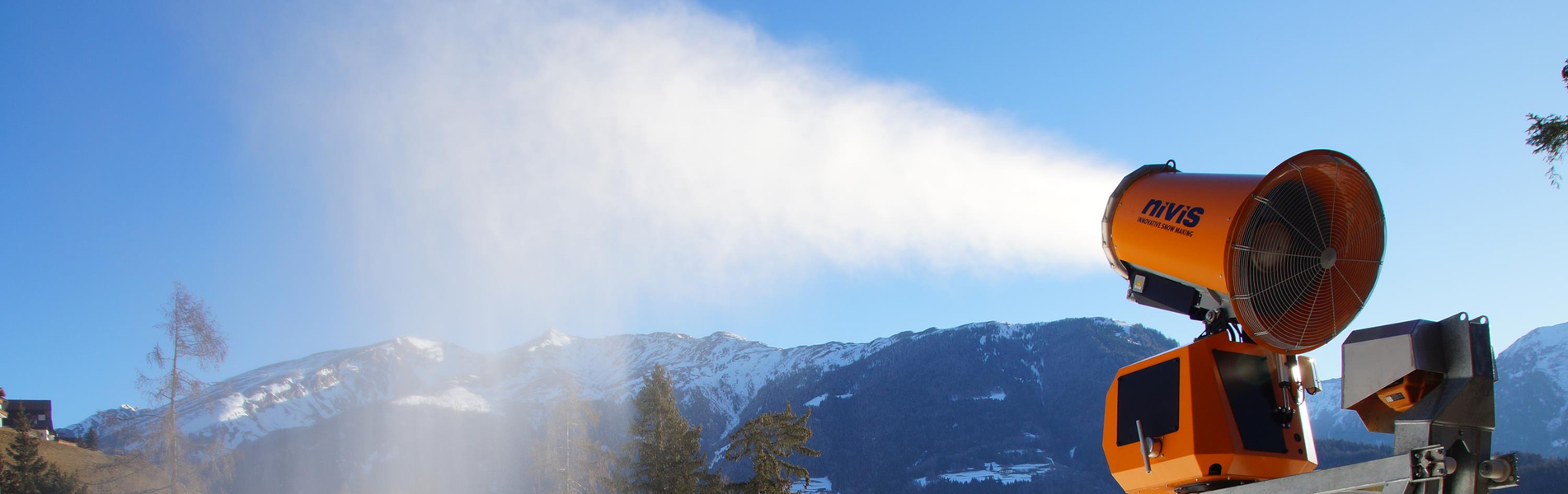 Orangefarbene Schneekanone erzeugt Schnee auf einer Skipiste vor Wald und Berglandschaft.
