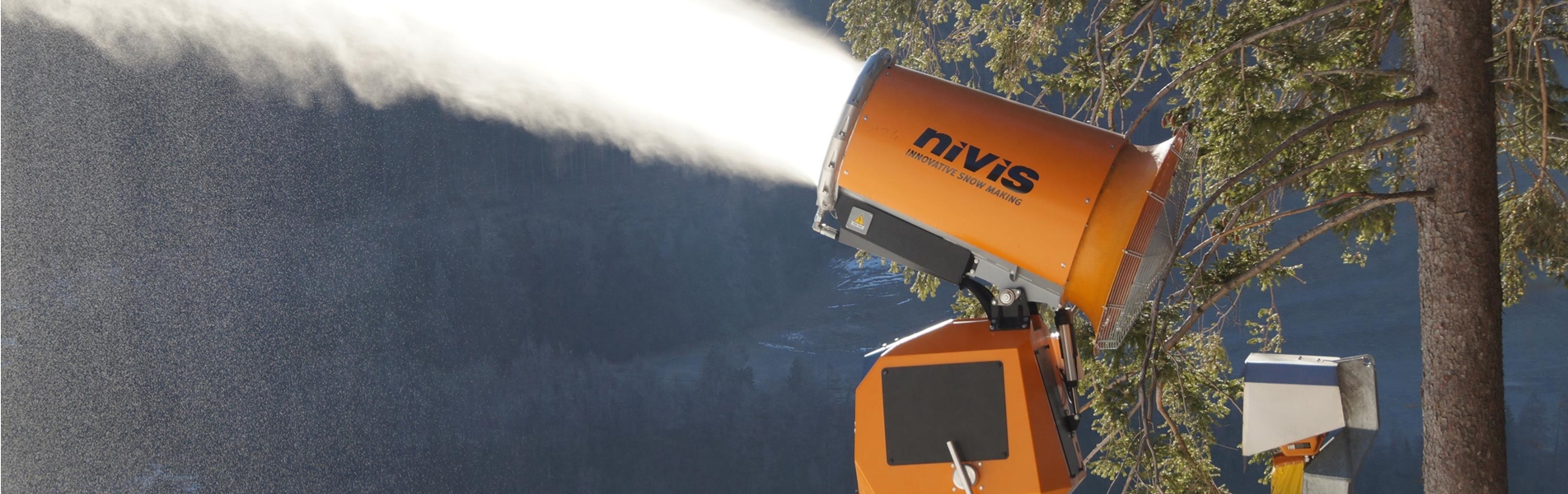 Orangefarbene Schneekanone versprüht Schnee vor bewaldeter Berglandschaft.